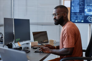 fotografia mostra um homem de óculos vestindo camiseta alaranjada sentado em frente aos computadores